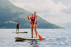 Two people in red life jackets are standing on the lake with their paddles on SUP boards, surrounded by green hills.