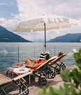 Two guests are lying on deckchairs under a white sunshade on a wooden walkway directly on the lake, enjoying the view.