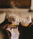 Two people are holding decorated whisky glasses from which light vapour is rising. The scene is bathed in warm, dim light.