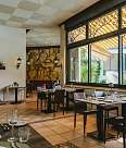 Spacious restaurant area with high windows overlooking a lush green terrace. The tables are elegantly set for guests