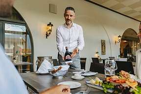 A hotel employee in a white shirt presents a bottle of red wine while a couple sits at the table looking forward to dinner