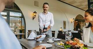Service personnalisé du vin à table Un employé de l'hôtel en chemise blanche présente une bouteille de vin rouge tandis qu'un couple est assis à la table en attendant le dîner