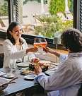 A couple toast with reddish-coloured drinks in a glass. On the table is a generous platter of appetisers, with palm trees and deckchairs in the background
