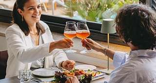 Apéritif élégant avec vue sur la campagne Un couple trinque avec des boissons de couleur rougeâtre dans un verre. Sur la table se trouve un généreux plateau d'entrées, avec des palmiers et des chaises longues en arrière-plan.