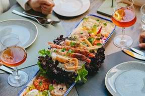 A rectangular serving dish with various seafood and salads. Next to it are plates, cutlery and glasses with a reddish-coloured drink.