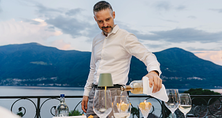 A hotel employee in a white shirt pours white wine into glasses on a terrace with a view of Lake Maggiore and the surrounding mountains.