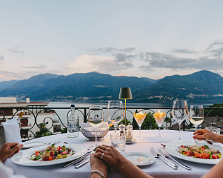 Un couple trinque avec des verres de vin, assis sur un quai du lac Majeur. En arrière-plan, les montagnes se dressent au coucher du soleil.