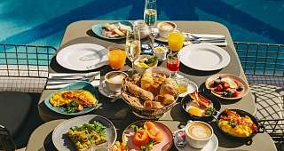 Petit-déjeuner varié au bord de la piscine Une table juste à côté de la piscine est richement dressée avec des omelettes, des sandwichs, du saumon, des fruits et diverses boissons, dont des jus de fruits et du café.