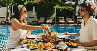 Déjeuner matinal au soleil au bord de la mer Un couple est assis au bord de la piscine et savoure un copieux petit-déjeuner composé de sandwichs, de fruits frais et de plats à base d'œufs, entouré de chaises longues et de parasols.