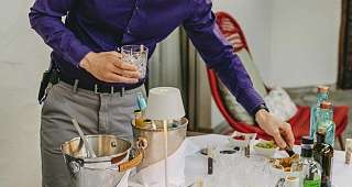 Dégustation de liqueurs avec les conseils d'experts Un employé de l'hôtel portant une chemise violette tient un verre transparent tout en présentant divers alcools sur une table avec des verres et des ingrédients