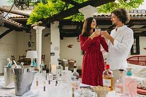 A couple clink glasses of gin in the courtyard. There are various bottles and spices on the table, with Mediterranean columns in the background