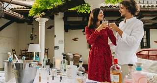Trinquez ensemble sur la terrasse Un couple trinque avec du gin dans la cour. Sur la table, il y a plusieurs bouteilles et des épices, avec des colonnes méditerranéennes en arrière-plan.