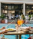 A couple are sitting at a table right by the pool. They are both enjoying their meal and toasting with orange aperitifs while chatting