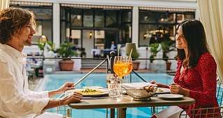 Dîner romantique au bord de la piscine Un couple est assis à une table juste à côté de la piscine. Tous deux apprécient leur repas et trinquent avec des apéritifs à l'orange tout en discutant.