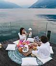 A couple are sitting at a festively decorated table on a terrace. In the background, the lake and mountains stretch out under an evening sky.