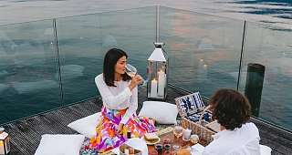 A couple are sitting at a festively decorated table on a terrace. In the background, the lake and mountains stretch out under an evening sky.
