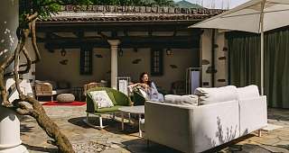 Détente dans le salon en plein air Une femme est assise, souriante, sur un fauteuil vert dans la cour de l'hôtel Sunstar de Brissago. Les meubles de la salle sont de couleurs vives et baignés de soleil.