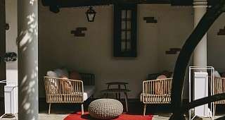 Coin salon confortable sous une pergola méditerranéenne Un coin couvert dans la cour intérieure avec des colonnes blanches, des poutres en bois et des fauteuils en rotin confortables. Un tapis rouge et un sol en pierre soulignent l'atmosphère méditerranéenne.