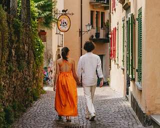 Un couple marche main dans la main le long d&#039;une ruelle pavée aux volets colorés dans un village méditerranéen au bord du lac Majeur.