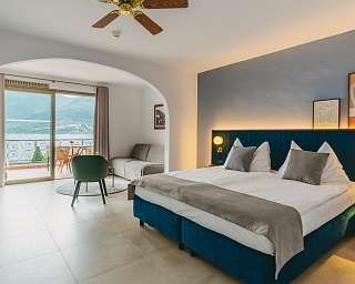 Chambre d&#039;hôtel lumineuse avec coin salon, télévision, fauteuil vert et vue directe sur le lac Majeur à travers de grandes portes-fenêtres