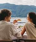 A couple sits happily at the breakfast table on the balcony. He holds a coffee cup in his hand while she laughs and enjoys her meal