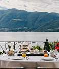 A table set with tiers full of dishes, juice glasses and a bouquet of flowers, directly above the lake with a view of the surrounding mountains
